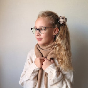 Könnte beinhalten: Eine junge Person mit langen blonden Haaren trägt einen weißen Pullover, einen braunen Schal und eine schwarze Brille. Sie trägt eine kleine Haarklammer mit Leopardenmuster.