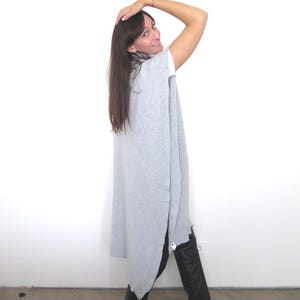 May include: A woman wearing a long, gray, sleeveless sweater with a high neckline and black pants. She is standing with her arm raised above her head, looking over her shoulder.