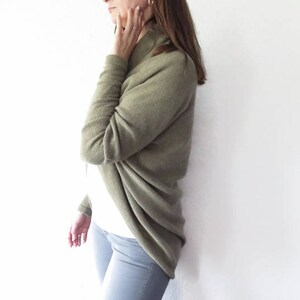 May include: A woman wearing a long-sleeved, olive green cardigan with a cowl neck. The cardigan is draped over her shoulders and is made of a soft, knitted fabric.