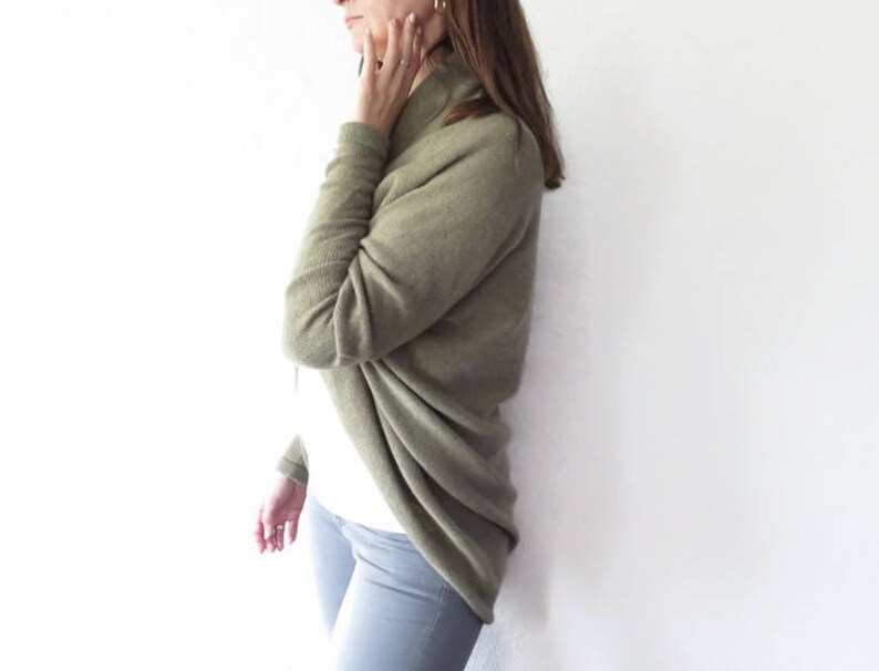 May include: A woman wearing a long-sleeved, olive green cardigan with a cowl neck. The cardigan is draped over her shoulders and is made of a soft, knitted fabric.