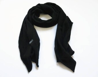 May include: A solid black scarf with a soft, matte texture and a small, rectangular grey fabric tag attached to one of the lower edges. The scarf is arranged in a loose, draped loop, showcasing its uniform dark color and simple, clean design.