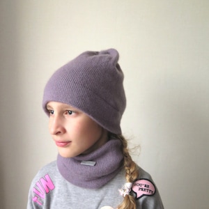 Può includere: Una giovane persona indossa un berretto di lana viola e un collo di lana abbinato. Il berretto è tirato giù sulla fronte. Indossa una felpa grigia con toppe rosa che dicono "You're Pretty" e "Love".