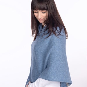 Pode incluir: Uma mulher vestindo um poncho de malha azul com uma camisa branca de manga comprida por baixo e cal&ccedil;as de ganga cinzas.