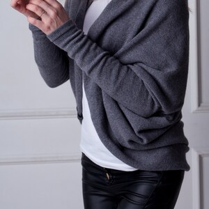 May include: A grey knit cardigan with a draped cowl neck and long sleeves. The cardigan is worn over a white t-shirt and black leather trousers.