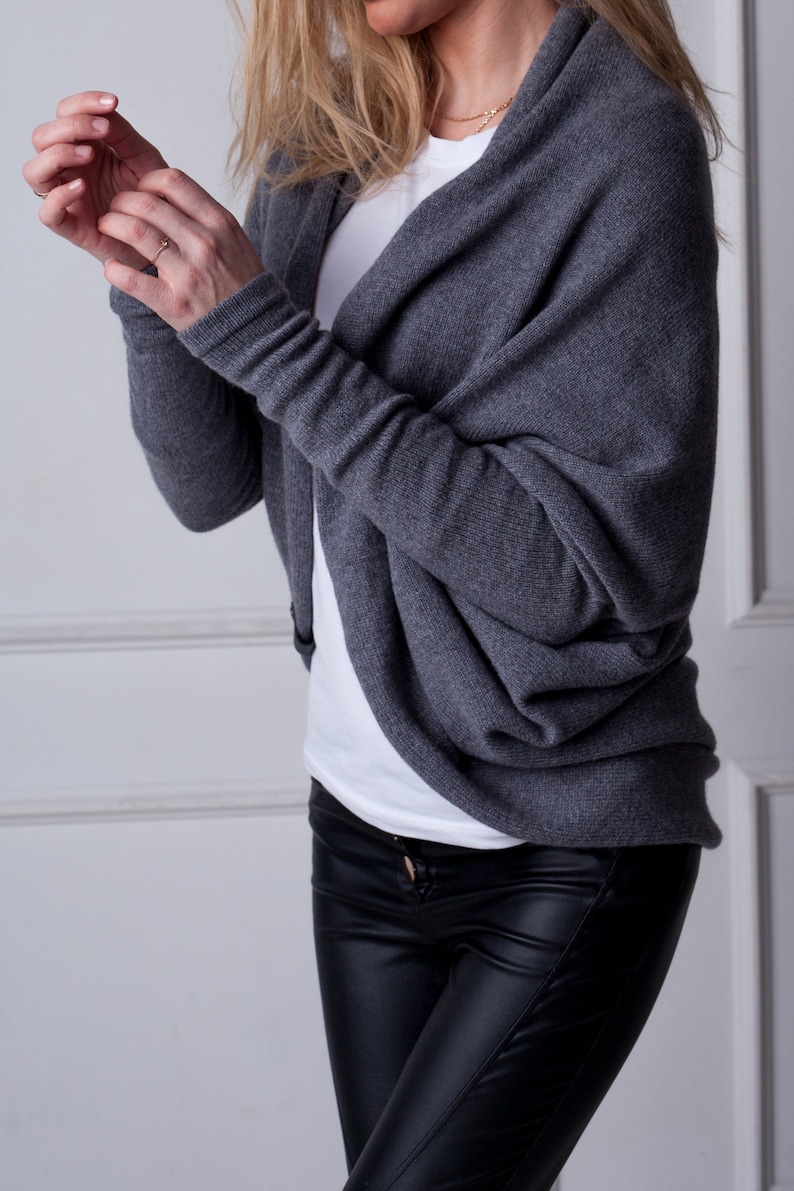 May include: A grey knit cardigan with a draped cowl neck and long sleeves. The cardigan is worn over a white t-shirt and black leather trousers.