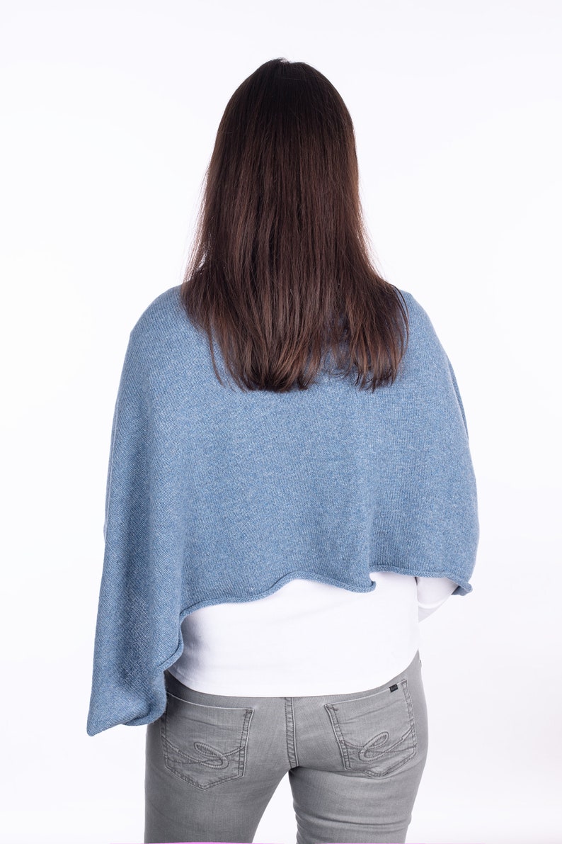 Pode incluir: Uma mulher vestindo um poncho de malha azul claro sobre uma camisa branca e cal&ccedil;as jeans cinzas.