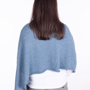 Pode incluir: Uma mulher vestindo um poncho de malha azul claro sobre uma camisa branca e cal&ccedil;as jeans cinzas.