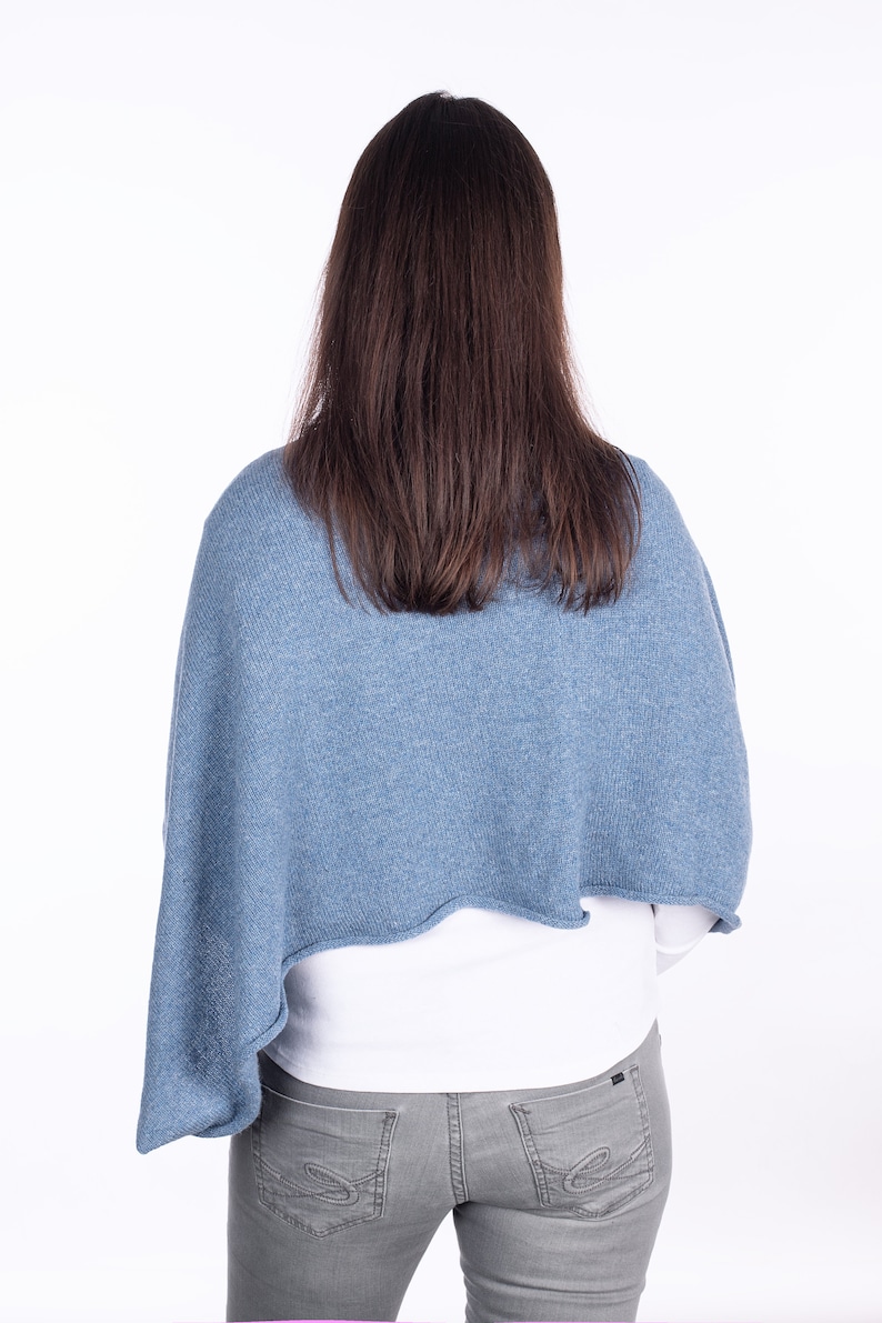 Pode incluir: Uma mulher vestindo um poncho de malha azul claro sobre uma camisa branca e cal&ccedil;as jeans cinzas.