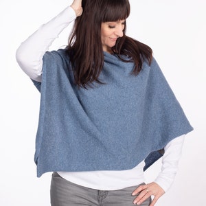 Pode incluir: Um poncho azul de malha com gola alta. O poncho &eacute; usado sobre uma camisa branca de manga comprida e cal&ccedil;as de ganga cinzas.