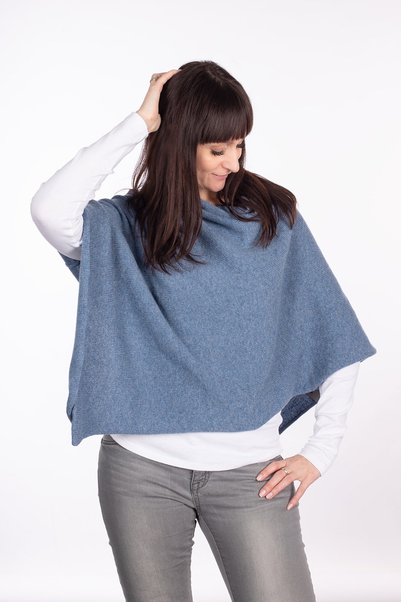 Pode incluir: Um poncho azul de malha com gola alta. O poncho &eacute; usado sobre uma camisa branca de manga comprida e cal&ccedil;as de ganga cinzas.