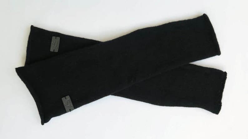 May include: A pair of black arm warmers with a gray logo on each. The arm warmers are made of a soft, stretchy fabric.