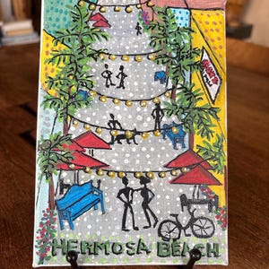 Puede incluir: Una pintura colorida de Hermosa Beach, con figuras, una bicicleta y sombrillas rojas. La obra de arte incluye luces de cadena, palmeras y el texto "Hermosa Beach" en la parte inferior. La pintura se exhibe sobre una superficie de madera.