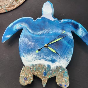 May include: A blue and white resin turtle clock with a wave design and gold accents. The clock face is surrounded by white foam waves and a sandy beach with seashells.