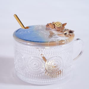 May include: A clear glass jar with a decorative pattern and a handle. The jar has a lid with a blue and white resin ocean scene and a small ceramic turtle figurine.
