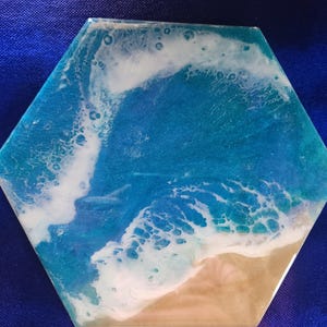 May include: A hexagonal resin art piece with a blue and white ocean wave design. The wave is crashing on a sandy beach.