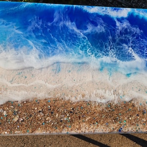 May include: A rectangular resin art piece depicting a beach scene. The top portion features a deep blue ocean with white foamy waves. The bottom section shows a sandy beach with embedded glitter and small stones.