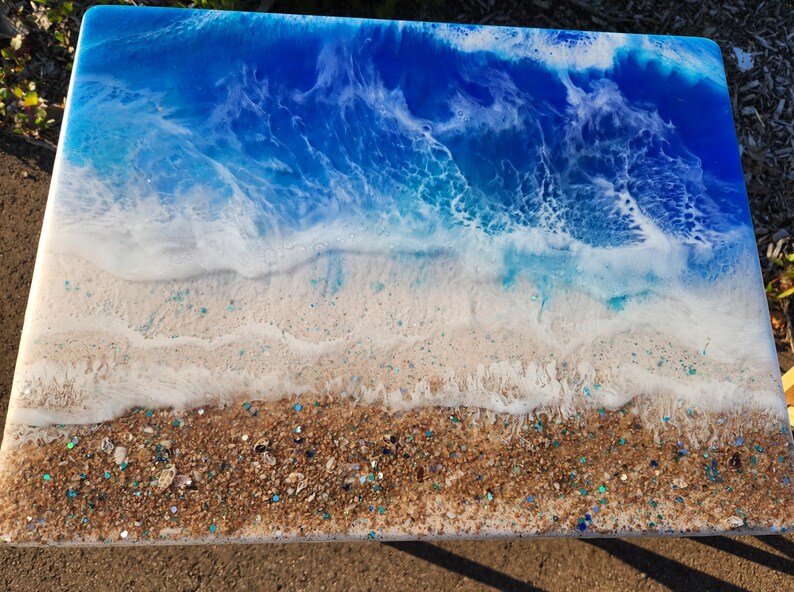 May include: A rectangular resin art piece depicting a beach scene. The top portion features a deep blue ocean with white foamy waves. The bottom section shows a sandy beach with embedded glitter and small stones.