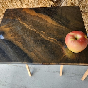 May include: A rectangular wooden table with a dark brown and gold marbled surface. The table has light-colored wooden legs. A red and yellow apple sits on the table. The table is approximately 24 inches long.