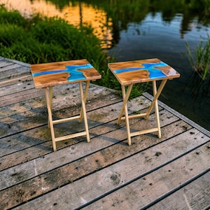 May include: Two rectangular folding tables featuring natural wood grain surfaces with a central inlay of bright blue resin. Each table is supported by a light-colored wooden frame with four legs that cross in an X-shape for stability.