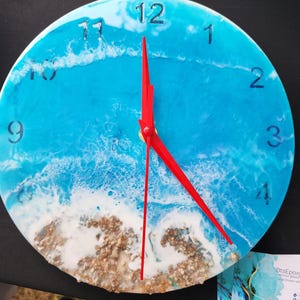 May include: Round wall clock with a blue and white ocean wave design. The clock face features black numerals and red hands. The bottom of the clock has a textured, sandy appearance.