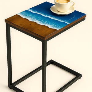 May include: A side table with a wooden top featuring a beach scene with blue waves and a sandy shore. The table has a black metal frame and a white coffee cup with a saucer on top. The table is 24 inches tall.