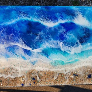 May include: A rectangular resin table top with a beach scene design. The top features shades of blue and white to mimic ocean waves, transitioning to a sandy beach with shells and small blue accents.
