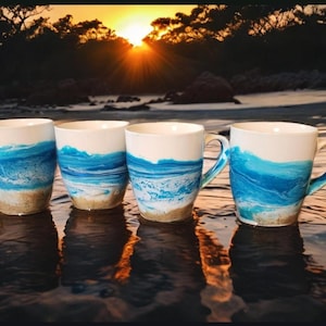 May include: Four ceramic mugs with a blue and white ocean wave design. The mugs are sitting on a wooden surface with a blurry background of a beach and sunset.
