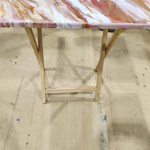 May include: A folding table with a rectangular top featuring a marbled design in shades of white, pink, and gold. The table has light-colored wooden legs. The table is designed for indoor use.