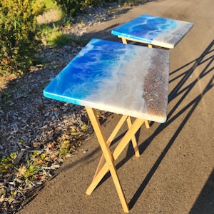 May include: Two wooden folding tables with blue and white resin tops depicting ocean waves and sand. The tables are set up on a gravel path.