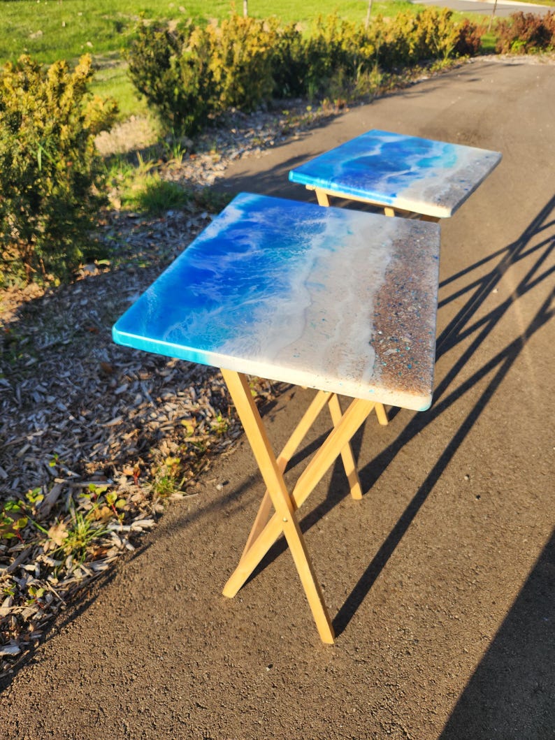 May include: Two square side tables with a beach-themed epoxy resin design. The tabletop features shades of blue, white, and tan, resembling ocean waves and sand. The legs are made of light-colored wood.