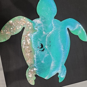 May include: A turquoise resin turtle-shaped wall art piece. The design incorporates a beach scene with white waves, sand, seashells, and black silhouettes of dolphins and birds. The artwork is on a black background.