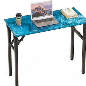 May include: A blue and white wave-patterned desk with black metal legs. A laptop, a coffee cup, and a stack of books are on the desk. The laptop screen displays a mountain landscape.