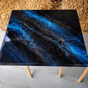 May include: Square table with a glossy black surface, swirled with blue and silver resin. The table has a modern design, supported by four wooden legs. The tabletop's design resembles a galaxy or ocean waves.