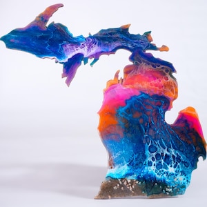 May include: A colorful abstract painting of the state of Michigan. The painting is done in shades of blue, purple, pink, orange, and white. The painting is on a wooden surface.