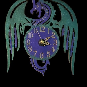 May include: A decorative wall clock featuring a dragon design. The clock has a blue face with gold numerals and hands. The dragon is teal and purple, with outstretched wings. The clock is mounted on a black background.