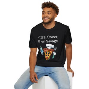 May include: A black t-shirt with a white graphic of a pizza slice wearing a chef's hat and holding a syringe and a fork. The text "Pizza: Sweet, then Savage" is printed on the shirt.