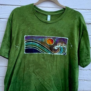 May include: Green t-shirt featuring a batik design of a wave and a sun. The design is within a rectangle and uses a dark blue, green, and orange colour scheme.