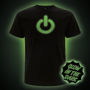 Puede incluir: Camiseta negra con un gráfico de botón de encendido que brilla en la oscuridad. El texto "Glow in the Dark" está en una pegatina en forma de estrella.