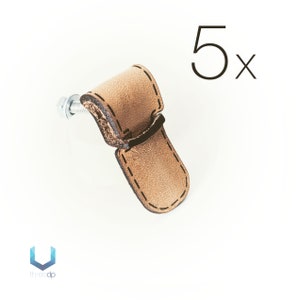 Pode incluir: Conjunto de cinco puxadores de armário de couro castanho com borda costurada e parafuso prateado. Os puxadores têm a forma de um retângulo curvo com borda arredondada.