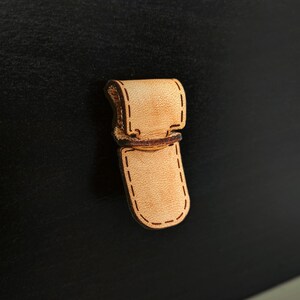 Pode incluir: Uma tira de couro marrom claro com costuras ao redor das bordas. A tira tem um laço em uma extremidade e uma aba na outra.