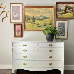 May include: White dresser with eight drawers and gold oval-shaped handles. The dresser has a curved bottom and sits on a rug in front of a wall with framed artwork.