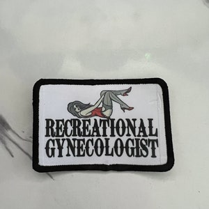 Op de afbeelding: Een witte patch met zwarte stiksels rond de randen. De patch heeft een zwart-witte illustratie van een vrouw in een rood-witte outfit. De tekst "RECREATIONAL GYNECOLOGIST" is in zwarte letters gedrukt.