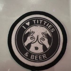 May include: A round, black and white patch with the text "I ❤️ TITTIES & BEER". The design features a heart symbol, two hands making an "okay" gesture, and a stylized image of breasts.