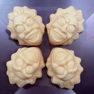 May include: Four small, beige, ceramic sculptures of faces with exaggerated features. The sculptures are arranged in a square pattern on a purple surface.