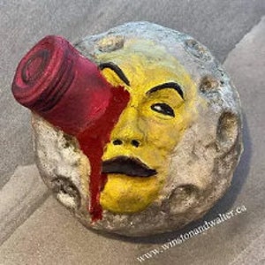 May include: A gray, moon-shaped sculpture with a human face painted on it. The face is yellow with a red, bloody eye and a red, bloody tear running down the cheek. A red, cylindrical object is attached to the sculpture's eye.