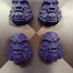 May include: Four purple 3D printed figurines of a creature with a scaly face and gills. The creature has a wide mouth with teeth and a furrowed brow.