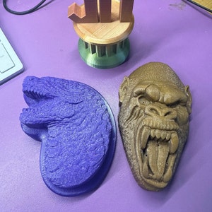May include: Two 3D printed figurines, one a purple crocodile head and the other a brown gorilla head with an open mouth, on a purple surface.