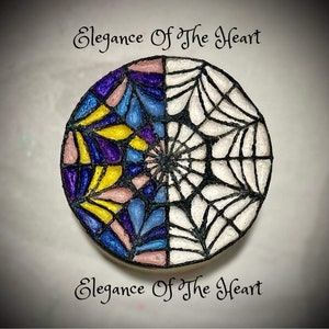 May include: A round, decorative object with a split design. One half features a stained glass pattern in shades of pink, yellow, blue, and purple. The other half is a white spiderweb design with black lines.