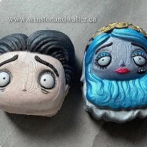 May include: Two bath bombs shaped like the heads of characters from the movie Corpse Bride. The bath bomb on the left is a pale pink color with black hair and large, white eyes. The bath bomb on the right is a blue color with long, blue hair and a white crown.