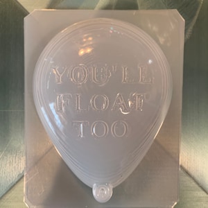 May include: A clear plastic mold shaped like a heart balloon with the text "You'll Float Too" inside. The mold is sitting on a gray surface with the text "Just Faerie Fizzies" below it.